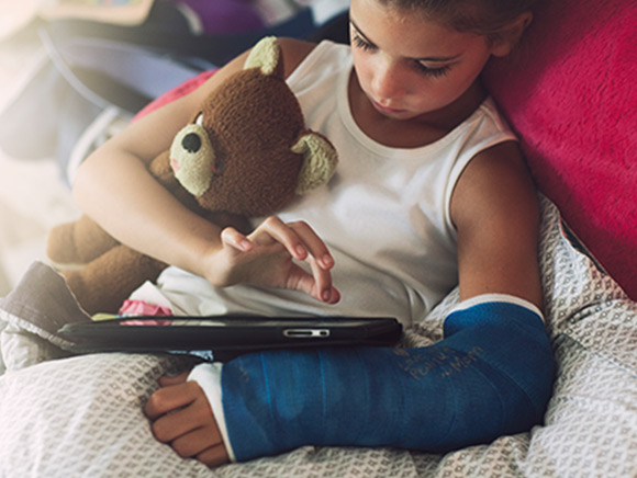 Little boy using a tablet with arm in a cast and teddy bear